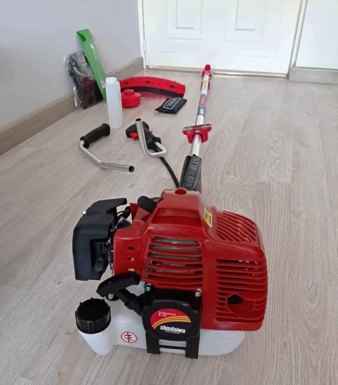 Guadaña motorizada roja y blanca desarmada sobre piso de madera, con motor, tubo, manubrios y diversos accesorios de ensamblaje.