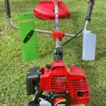 Guadaña motorizada roja con mango ergonómico, protector, tanque blanco, botella de combustible y empaque de cuchilla sobre césped verde.