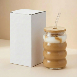 Vaso ondulado de vidrio con tapa de bambú, pitillo y caja blanca sobre superficie beige.