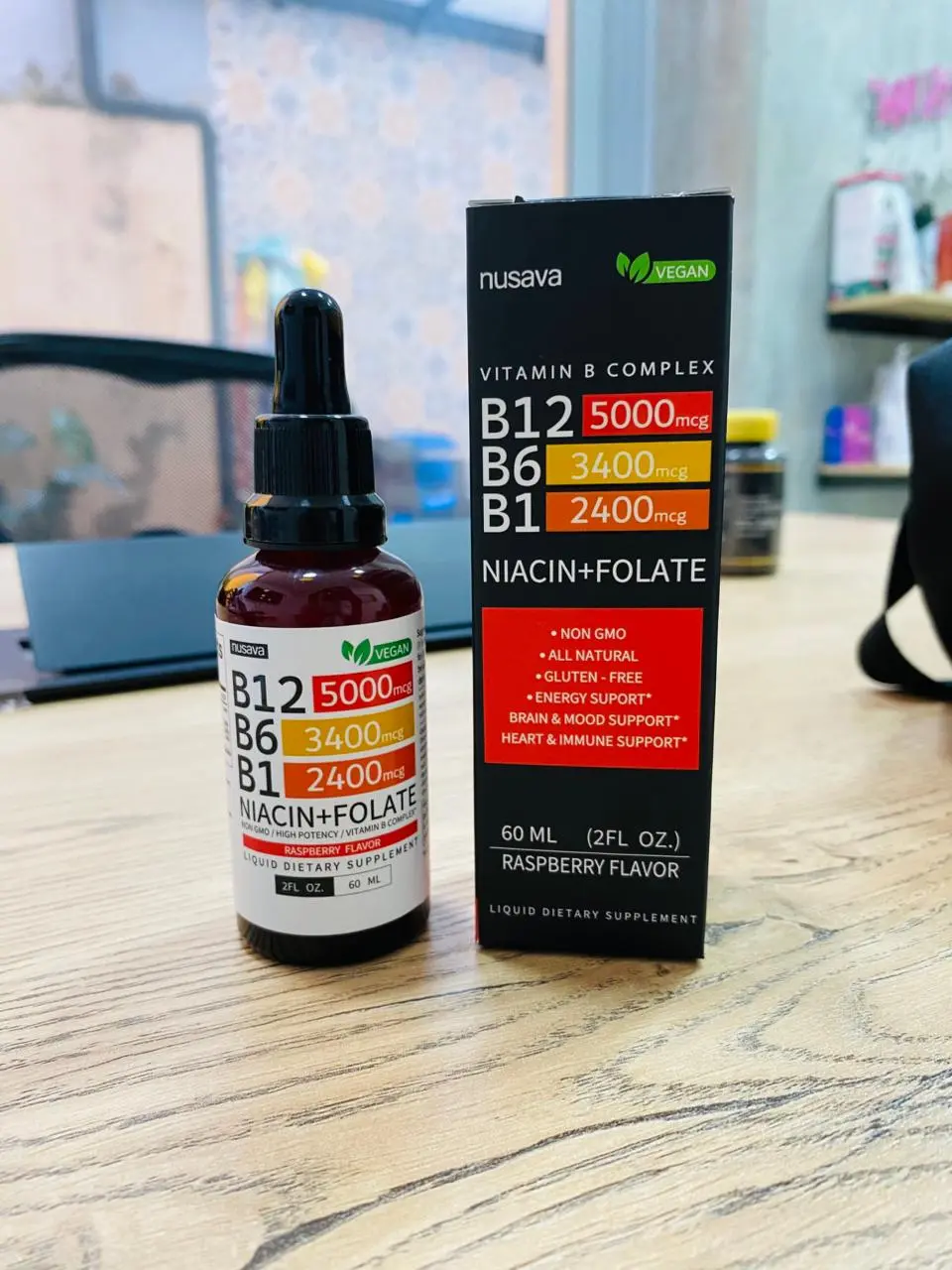 Frasco gotero de vidrio ámbar con etiqueta y caja negra de Nusava Vitamina B Complex 60 ml con frambuesa sobre mesa de madera.