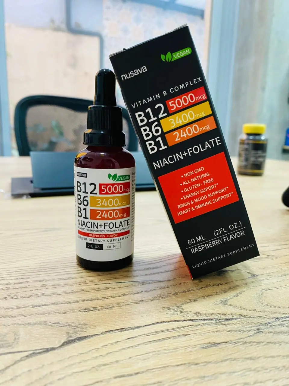 Gotero de vitamina B12 líquida junto a su caja negra en una mesa de madera, con presentación de 60 ml y sabor frambuesa.