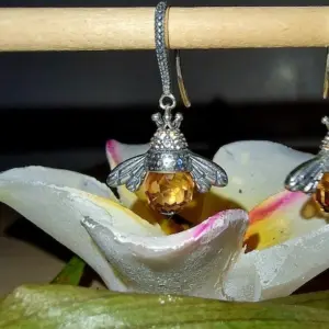 Pendientes de abeja vintage en plata con cristales ámbar colgando de una barra de madera sobre flor blanca.
