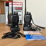 Dos radios portátiles Baofeng negros con antena larga sobre bases de carga, acompañados de caja, manual y accesorios en mesa de madera.