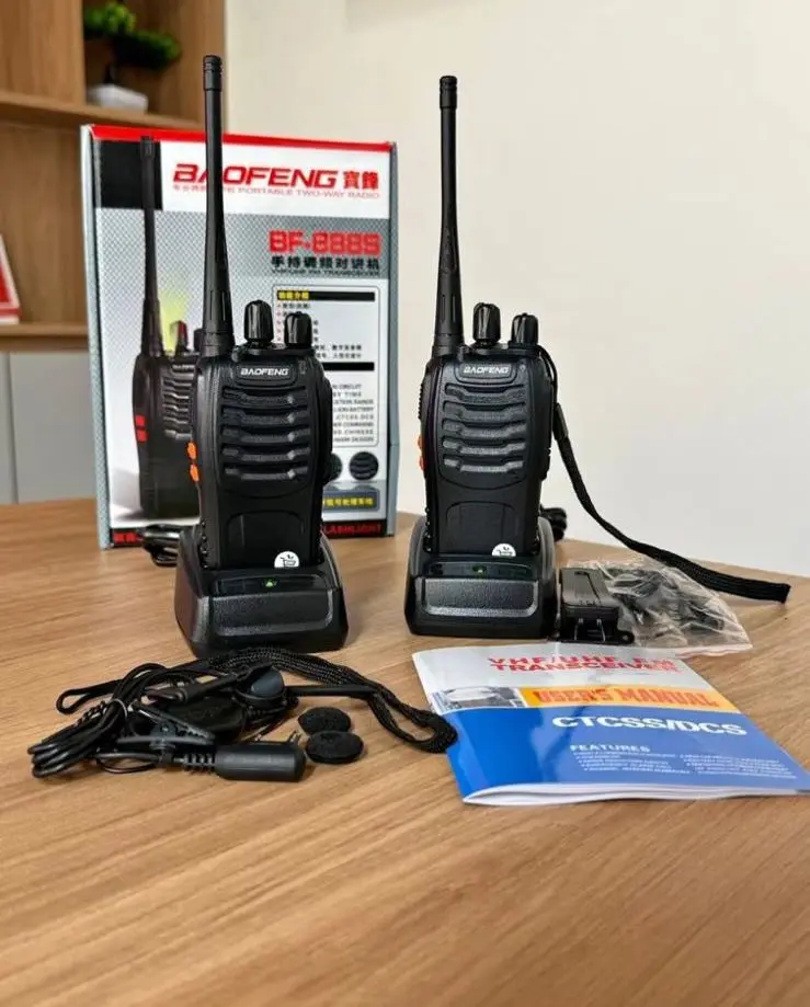 Dos radios portátiles Baofeng negros con antena larga sobre bases de carga, acompañados de caja, manual y accesorios en mesa de madera.