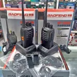 Par de radios portátiles Baofeng negros en sus bases de carga sobre cajas, con dos audífonos y accesorios en bolsas plásticas.