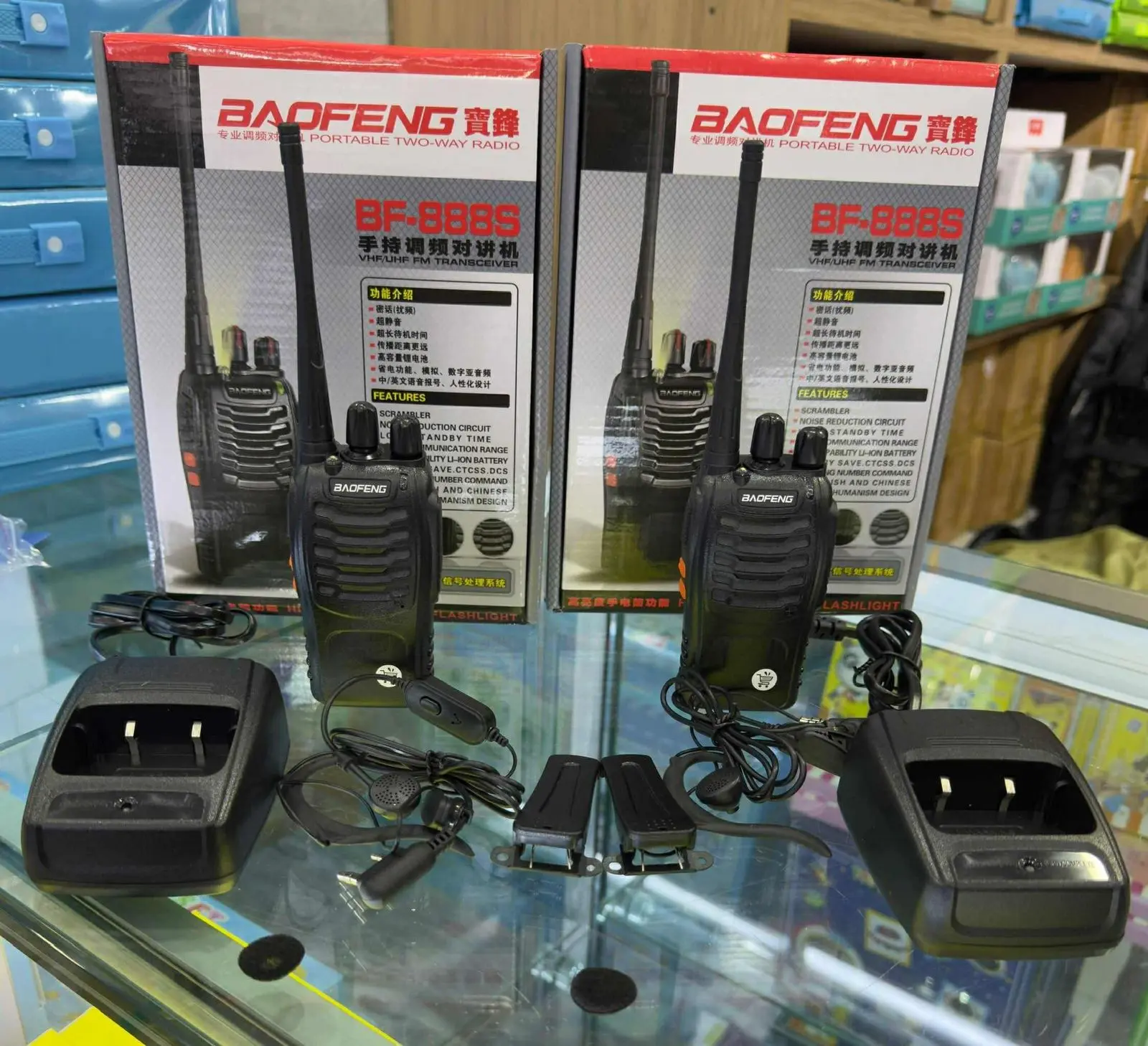 Dos radios portátiles Baofeng BF-888S negros con antenas, sobre cajas de empaque, acompañados de bases de carga, manos libres y clips.