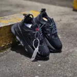 Zapatos Versace negros de diseño moderno con suela robusta y textura de malla, apoyados sobre un bordillo amarillo en un entorno urbano.