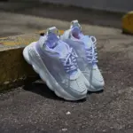 Tenis Versace blancos y lavanda con suela gruesa ondulada, apoyados en un bordillo amarillo en un entorno urbano.