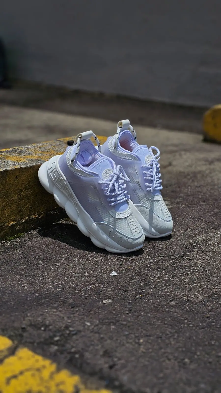Tenis Versace blancos y lavanda con suela gruesa ondulada, apoyados en un bordillo amarillo en un entorno urbano.