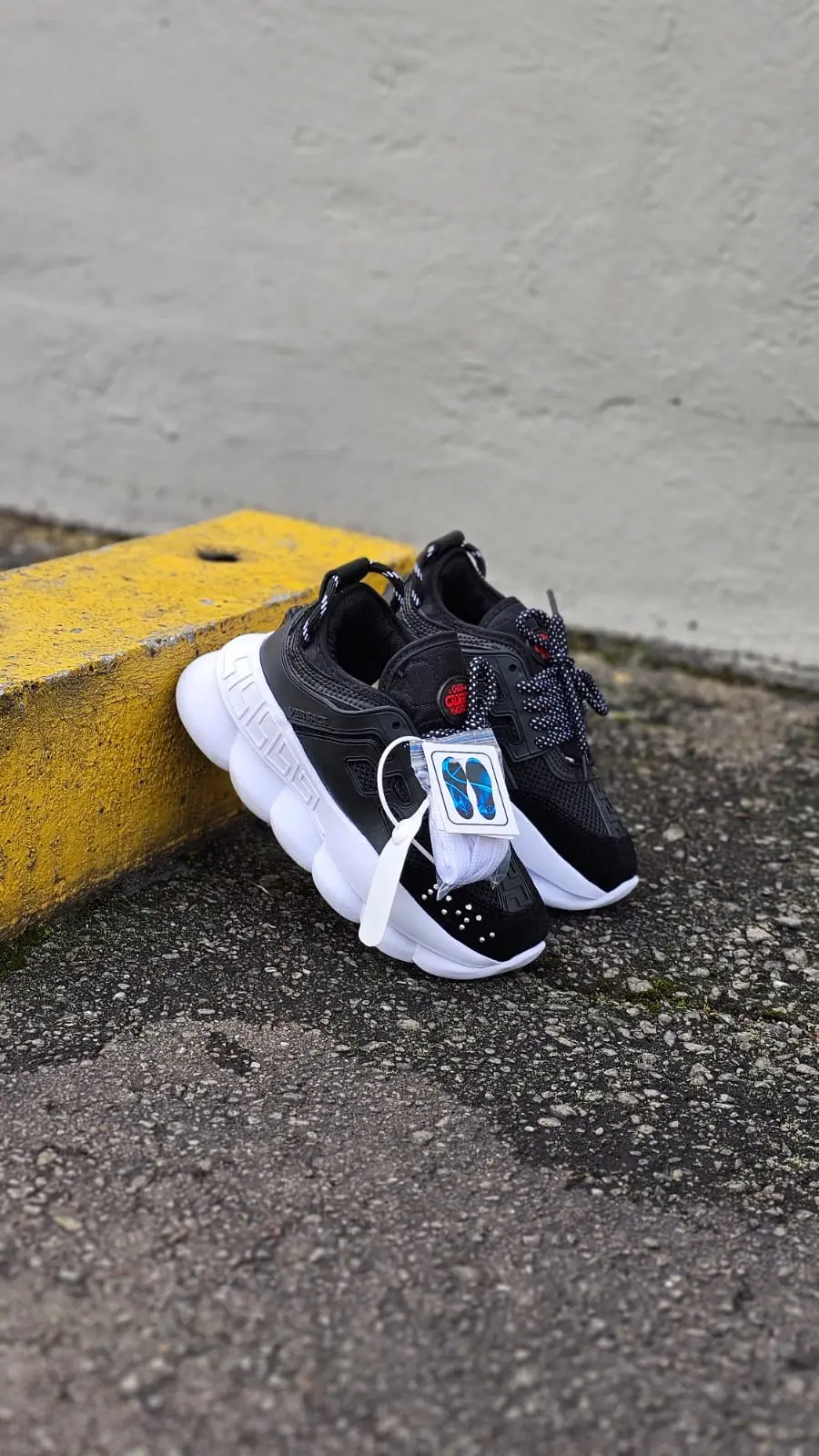 Zapatos Versace negros de diseño moderno con suela blanca gruesa y detalles rojos, apoyados en un bordillo amarillo.