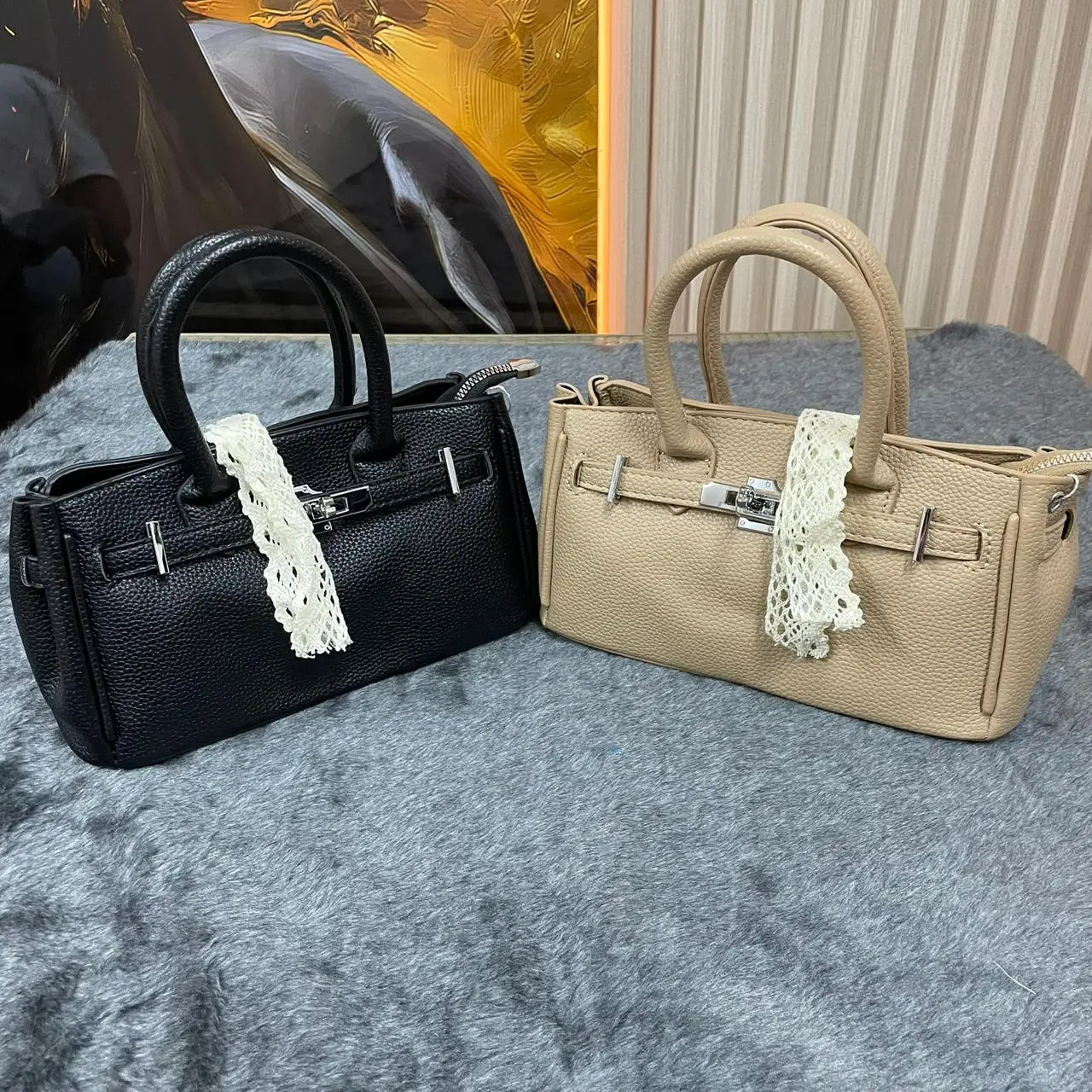 Dos bolsos semi-rígidos para dama en negro y beige con textura granulada, asas cortas y detalle decorativo de encaje blanco.