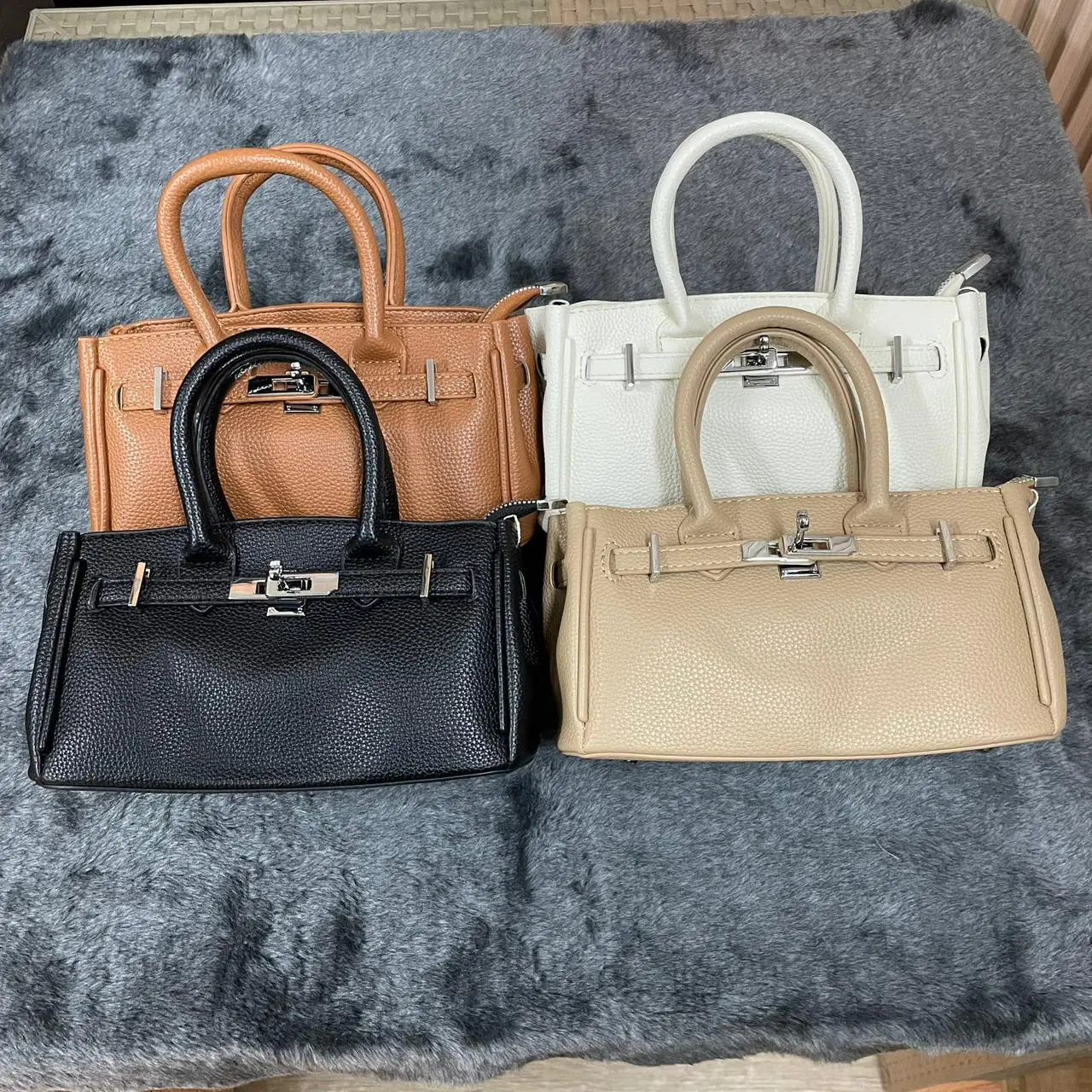 Cuatro bolsos semi-rígidos para dama en colores negro, beige, blanco y camel con cierres metálicos sobre fondo gris texturizado.