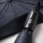 Sombrilla automática compacta negra con mango de botón y tela resistente, vista en ángulo para resaltar su mecanismo y textura.