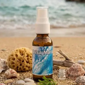 Spray aromático Brisa Marina en frasco ámbar con etiqueta de olas, rodeado de conchas, esponja y madera sobre arena en un entorno costero.