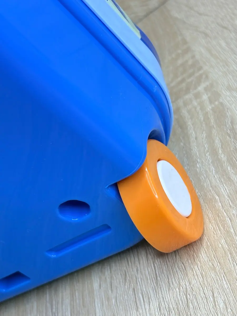 Rueda naranja con centro blanco de maleta de juguete azul sobre piso de madera.