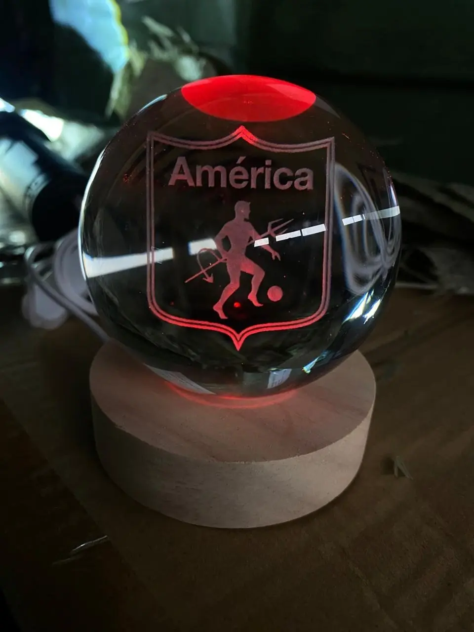 Lámpara LED esférica de cristal con grabado láser del escudo del América de Cali, iluminada en rojo sobre base de madera circular.