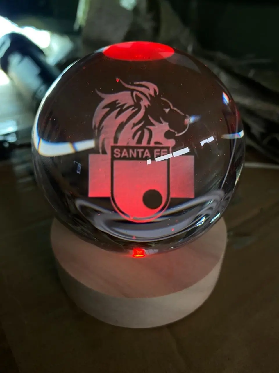 Lámpara LED esférica de cristal con grabado láser del escudo de Santa Fe, iluminada en color rojo sobre base de madera circular.