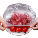 Manos estirando una tapa elástica transparente sobre un plato con tomates rojos, demostrando su función para conservar alimentos frescos.
