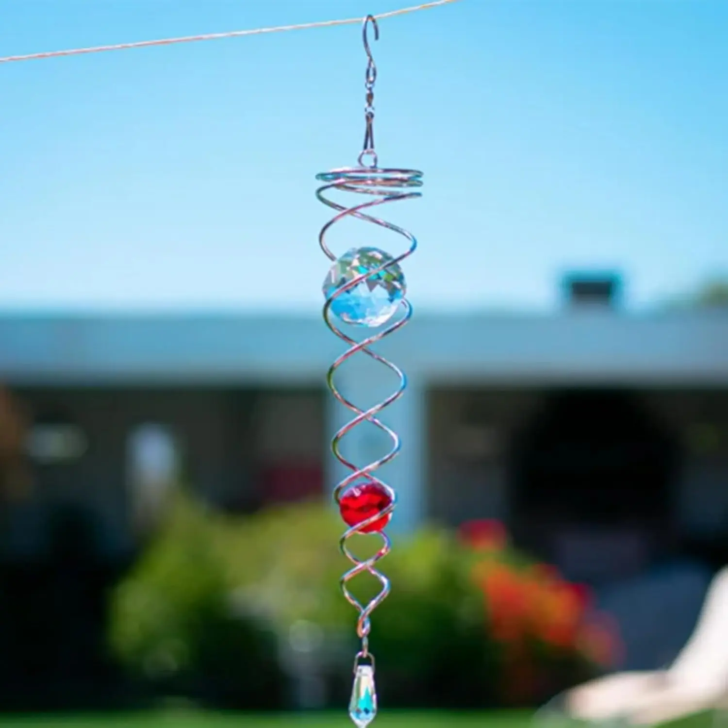 Espiral colgante de metal plateado con una bola de cristal azul arriba y una roja abajo, suspendida en el jardín bajo el sol.