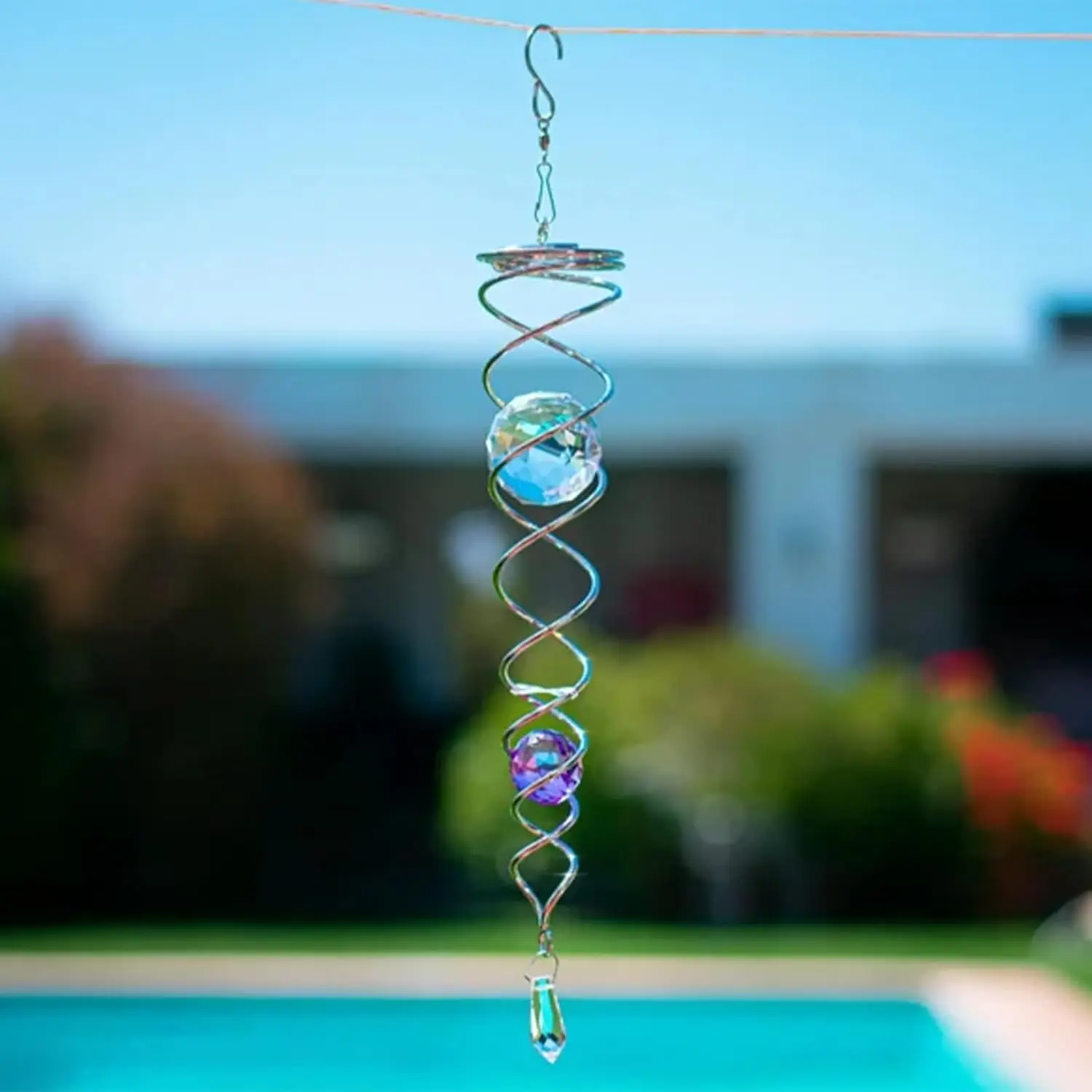 Espiral decorativa metálica colgante para jardín con dos esferas de cristal facetadas en tonos azul y morado y un prisma inferior, suspendida al aire libre frente a una piscina.