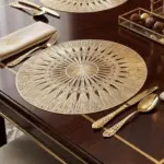 Individual redondo de PVC sintético dorado con diseño de radios sobre mesa de madera oscura con cubiertos y servilleta.