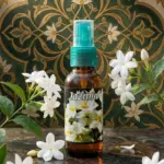 Atomizador ámbar de 60 ml con fragancia de jazmín natural, sobre superficie de mármol con flores blancas y fondo ornamental verde y dorado.