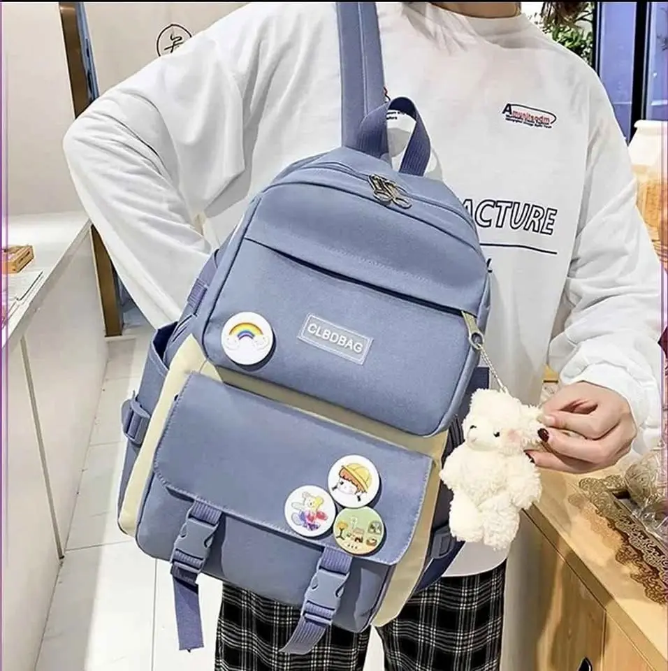 Morral escolar azul de tela Oxford con múltiples compartimentos, decorado con pines y un llavero de peluche blanco, mostrado en uso.