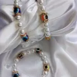 Dos pulseras de perlas sintéticas blancas con cuentas de ojo turco en varios colores y separadores dorados con brillantes sobre tela de satén blanca.