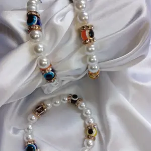 Dos pulseras de perlas sintéticas blancas con cuentas de ojo turco en varios colores y separadores dorados con brillantes sobre tela de satén blanca.