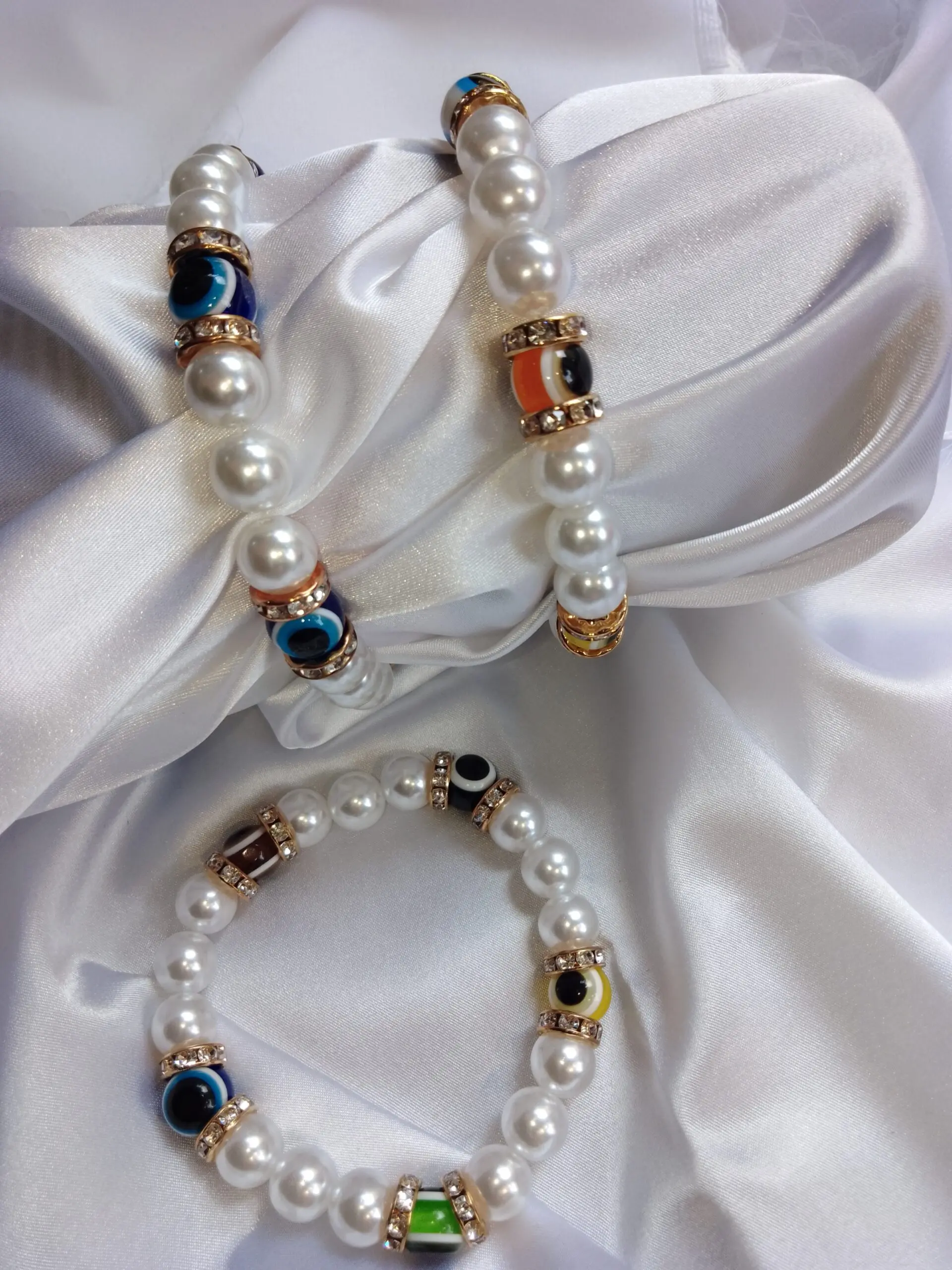 Dos pulseras de perlas sintéticas blancas con cuentas de ojo turco en varios colores y separadores dorados con brillantes sobre tela de satén blanca.