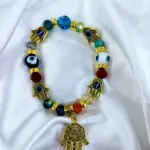 Pulsera elástica dorada con cuentas de vidrio multicolor y dijes de mano de Fátima y ojo turco, sobre fondo de tela blanca brillante.