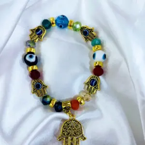 Pulsera elástica dorada con cuentas de vidrio multicolor y dijes de mano de Fátima y ojo turco, sobre fondo de tela blanca brillante.