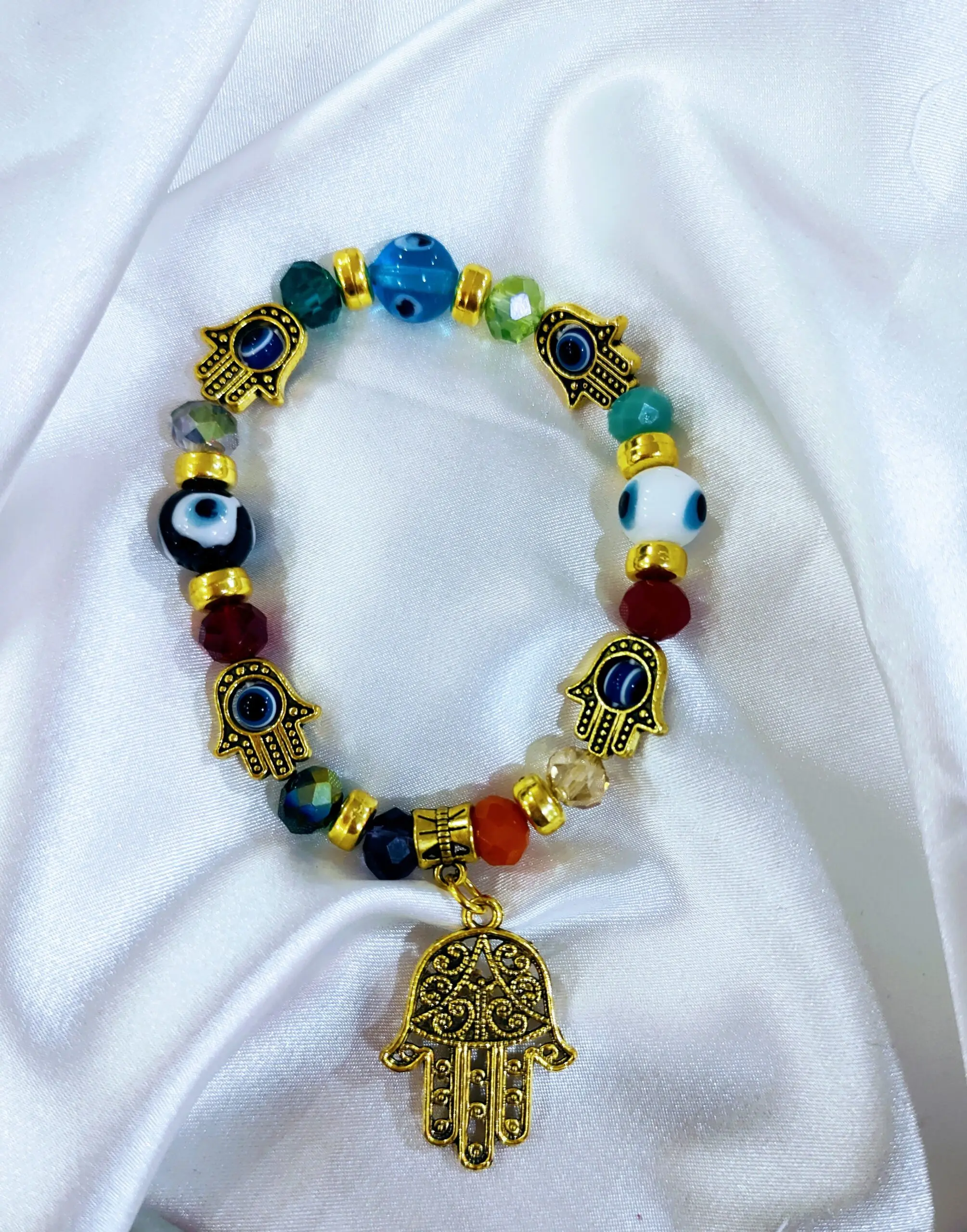 Pulsera elástica dorada con cuentas de vidrio multicolor y dijes de mano de Fátima y ojo turco, sobre fondo de tela blanca brillante.
