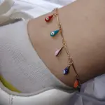 Tobillera dorada con dijes colgantes en forma de gota multicolor y diseño de ojo turco, usada sobre calcetín blanco y zapatilla.