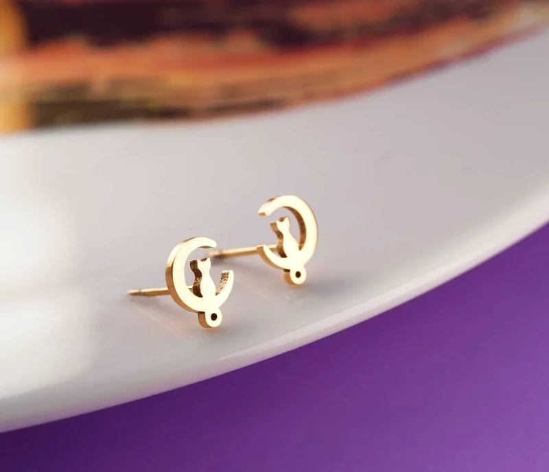 Aretes topitos dorados de acero inoxidable con diseño de luna creciente y silueta de gato, mostrados en primer plano sobre fondo blanco.