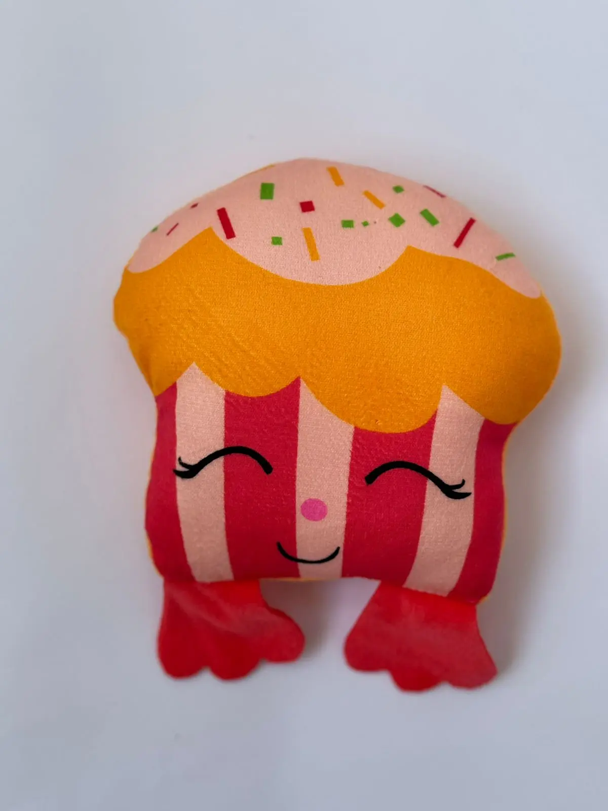 Peluche de cupcake para perro con cara sonriente, chispas de colores, cuerpo de rayas rojas y rosadas y pequeños pies rojos.
