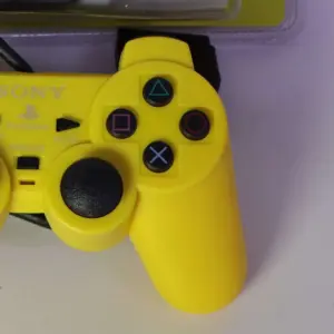 Control para videojuegos tipo PlayStation 2 alámbrico en color amarillo brillante con joysticks negros, sobre superficie blanca.