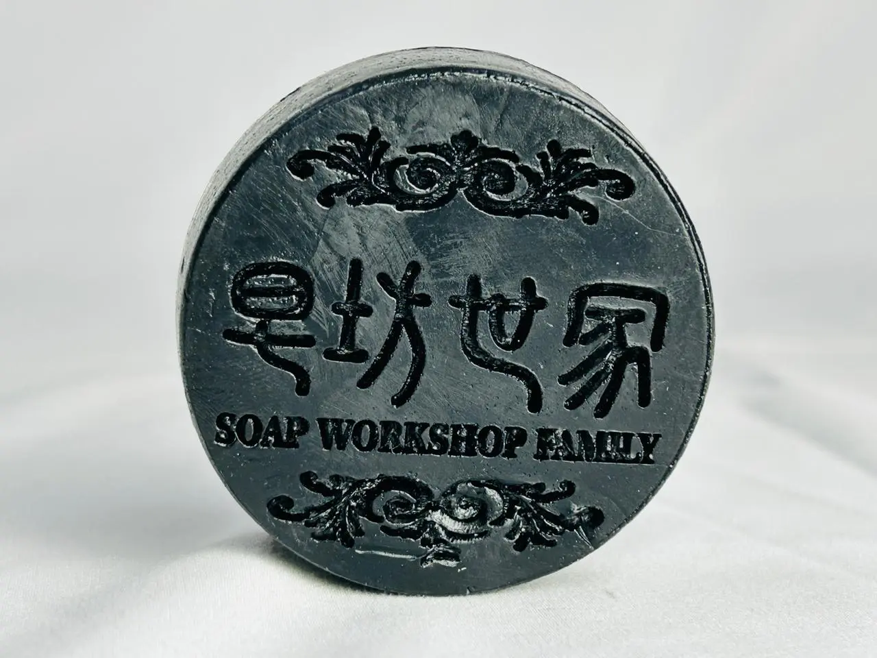 Shampoo en barra redondo color negro, con relieve y texto en chino y en inglés "Soap Workshop Family", sobre fondo blanco.