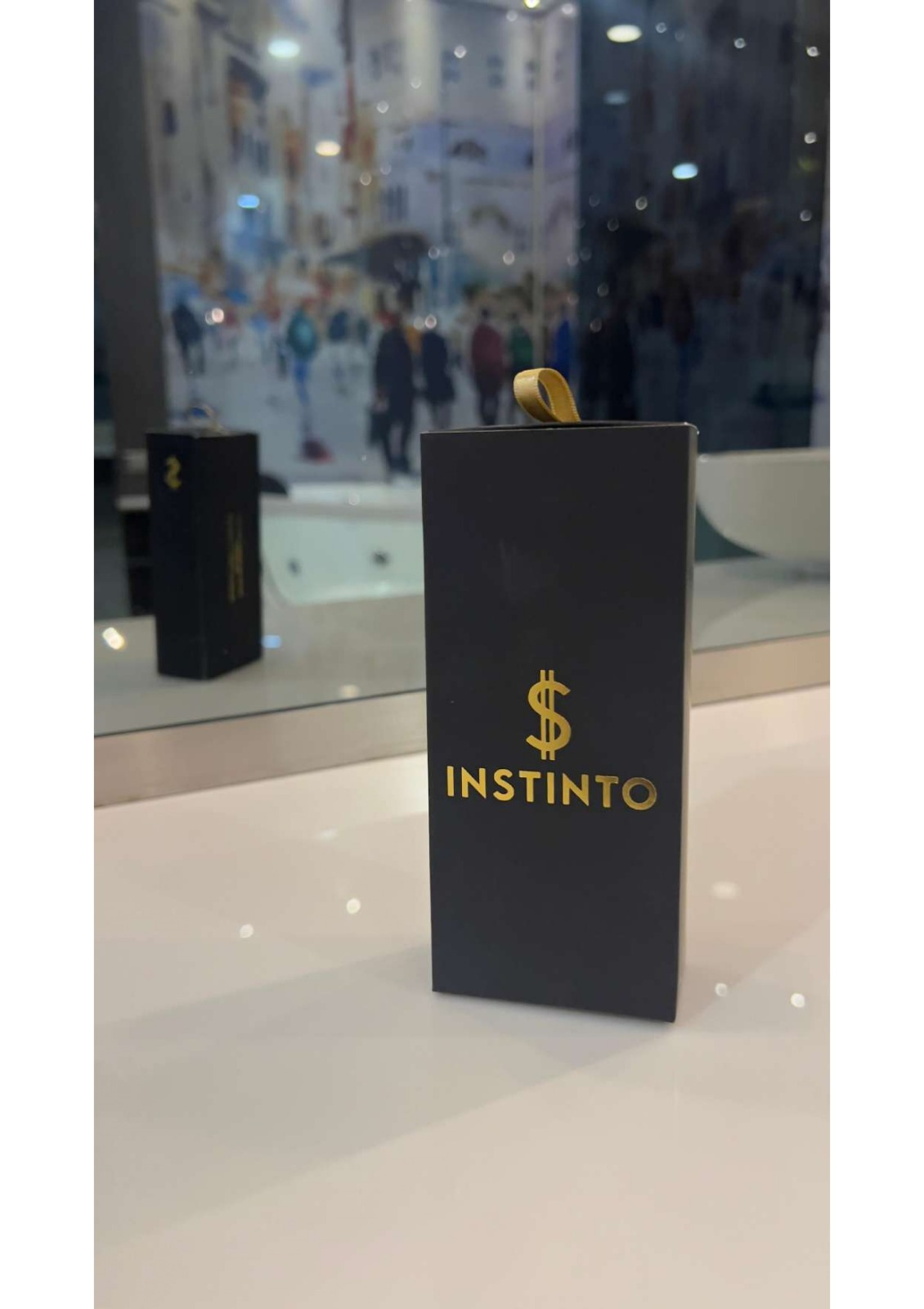 Caja negra rectangular de perfume Instinto con logo dorado de signo pesos, ubicada sobre superficie blanca brillante con reflejo en espejo al fondo.