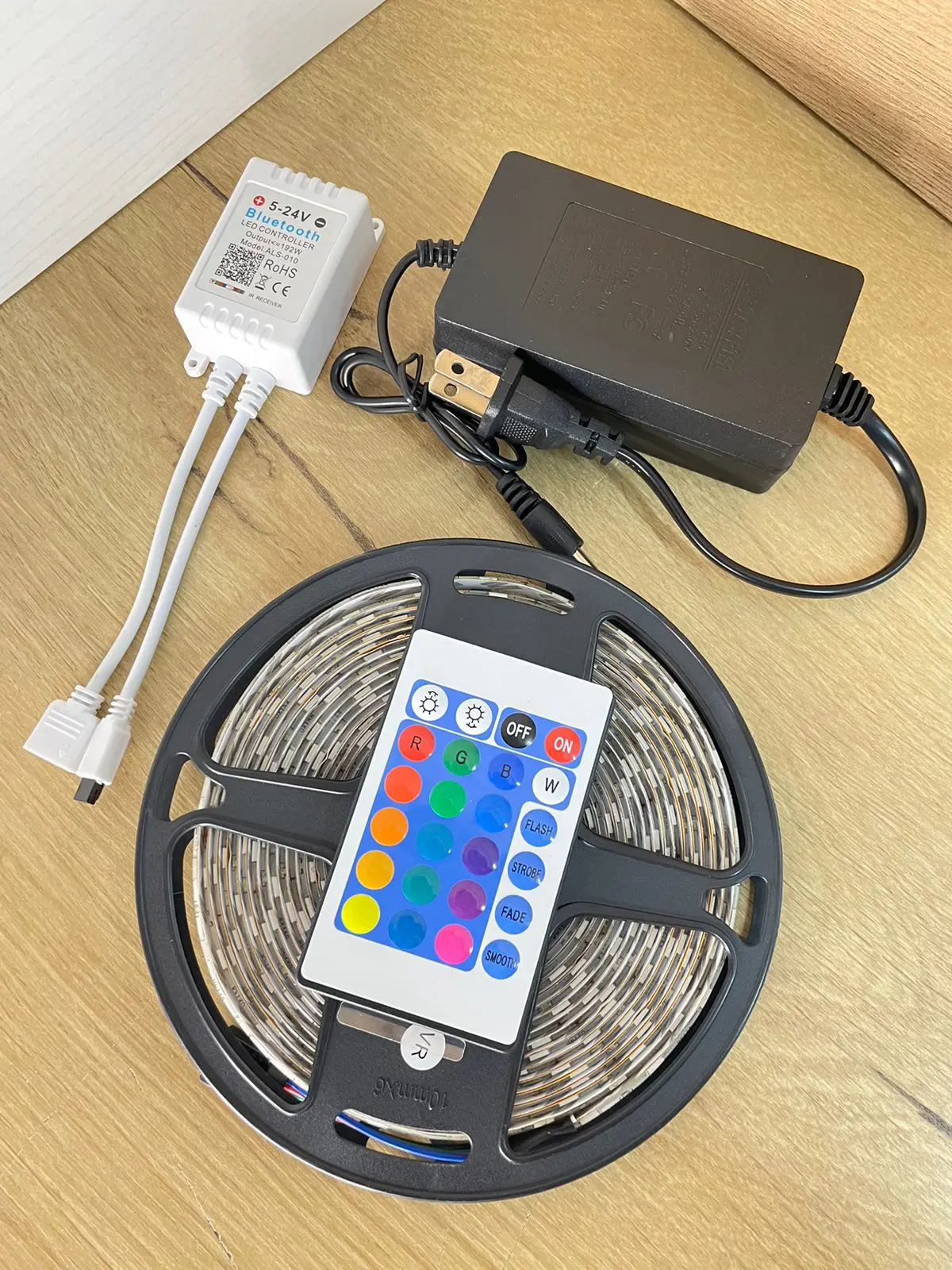 Kit de cinta LED RGB de 5 metros con controlador Bluetooth blanco, control remoto multicolor, fuente de alimentación y cables conectores sobre madera.