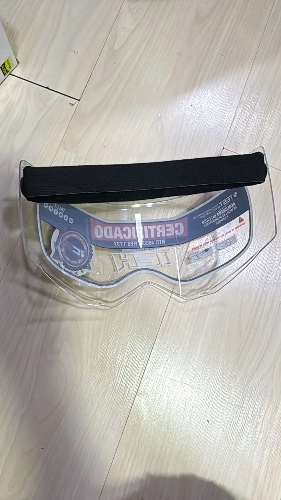 Visor transparente de policarbonato para casco con banda elástica negra superior, colocado sobre piso de madera.