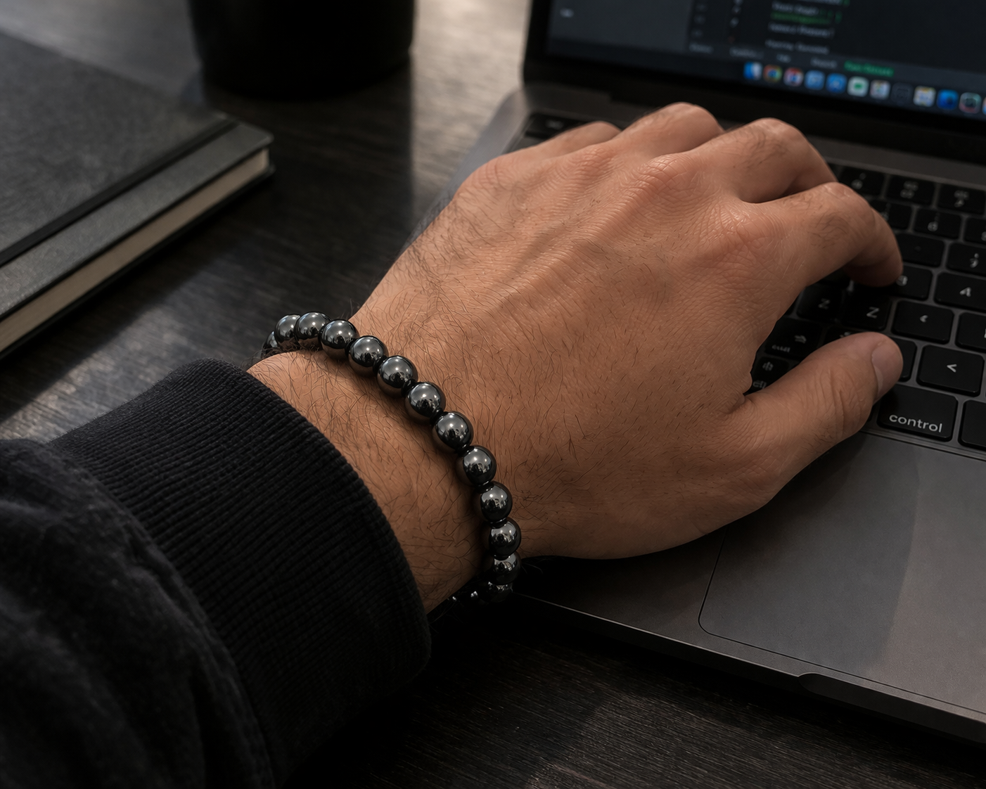 Pulsera de hematita natural con cuentas esféricas gris metalizado en la muñeca de una persona usando un computador portátil.