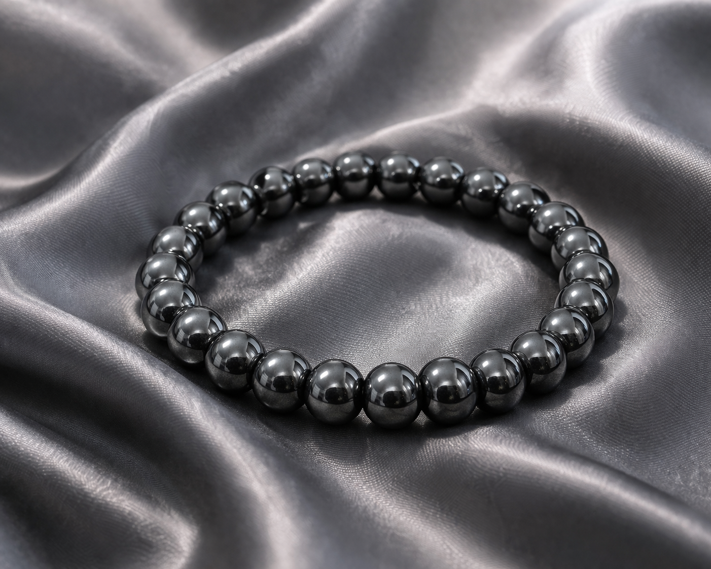Pulsera de hematita natural con cuentas esféricas de color gris metálico brillante sobre tela satinada gris.