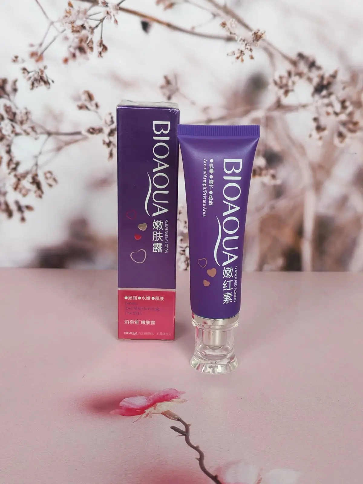 Crema aclaradora íntima Bioaqua en tubo morado con dispensador transparente, acompañada de su caja vertical sobre fondo rosa con flores desenfocadas.