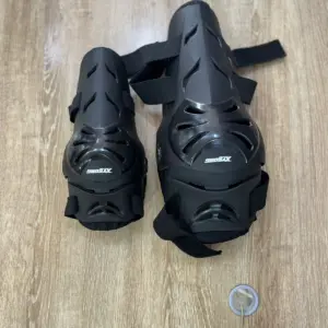 Par de protecciones negras para moto de alto impacto, una rodillera grande y una codillera más pequeña con ventilación y correas ajustables sobre madera.