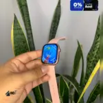 Smartwatch rosa con pantalla AMOLED y correa texturizada, sostenido en mano frente a planta de hojas largas.