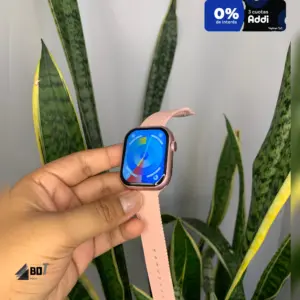 Smartwatch rosa con pantalla AMOLED y correa texturizada, sostenido en mano frente a planta de hojas largas.