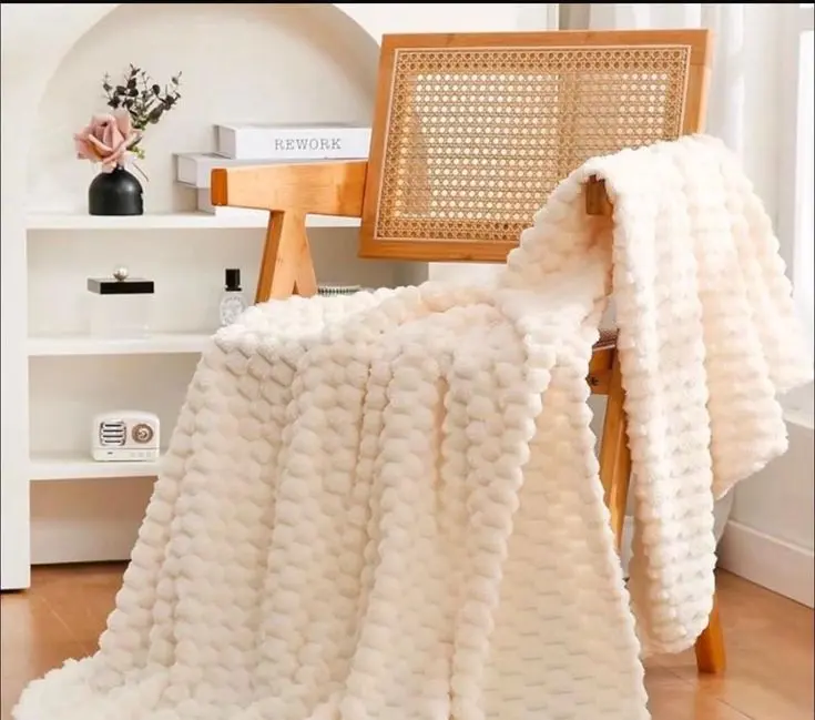 Cobija de textura tortuga color crema con relieve suave, extendida sobre una silla de madera en un ambiente decorado y acogedor.