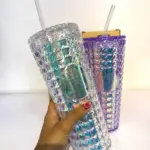 Mano sosteniendo vaso Starbucks reutilizable tornasolado con textura de rombos, tapa y pitillo, junto a otro vaso morado de fondo.