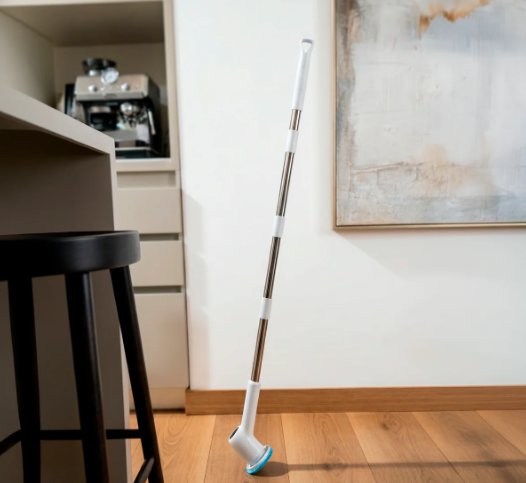 Cepillo eléctrico giratorio inalámbrico blanco con mango largo metálico apoyado sobre piso de madera en una sala.